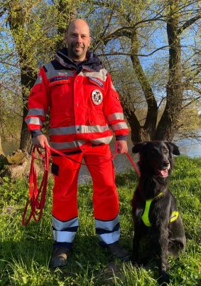 Ein Rettungskräfte in roter Uniform steht mit einem schwarzen Spürhund vor Bäumen.
