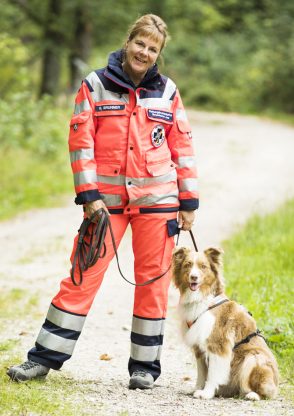 Frau in orangefarbener Einsatzkleidung mit einem ehrenamtlichen Rettungshund auf einem Weg im Wald.