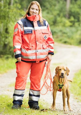 Frau in orangefarbener Einsatzkleidung steht mit Mantrailing Hund an einem Waldweg.