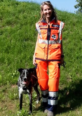 Frau in orangefarbener Einsatzkleidung mit einem Rettungshund auf einer Wiese.