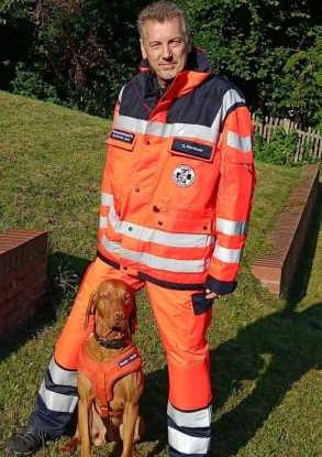 Mann in orange Rettungsanzug steht neben einem Rettungshund, ebenfalls in orange.