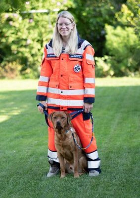 Frau in orangefarbener Einsatzkleidung steht mit einem Mantrailinghund auf einer Wiese.