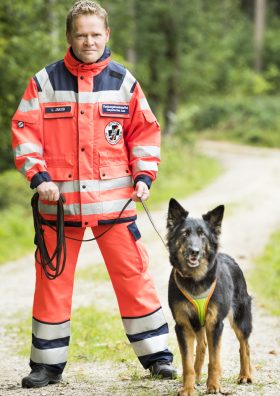 Ein Mantrailer in orangefarbener Uniform steht neben einem Rettungshund.