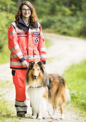 Person in roter Einsatzbekleidung steht neben einem Collie (Suchhund) auf einem Waldweg.