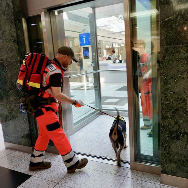 Ein Rettungshundeführer in orangefarbener Uniform führt einen Spürhund in einen Aufzug.