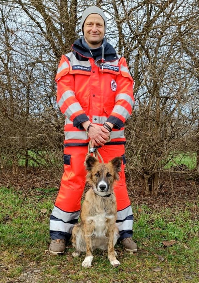 Ein Rettungshundeführer in orangefarbener Einsatzkleidung steht neben einem Rettungshund.