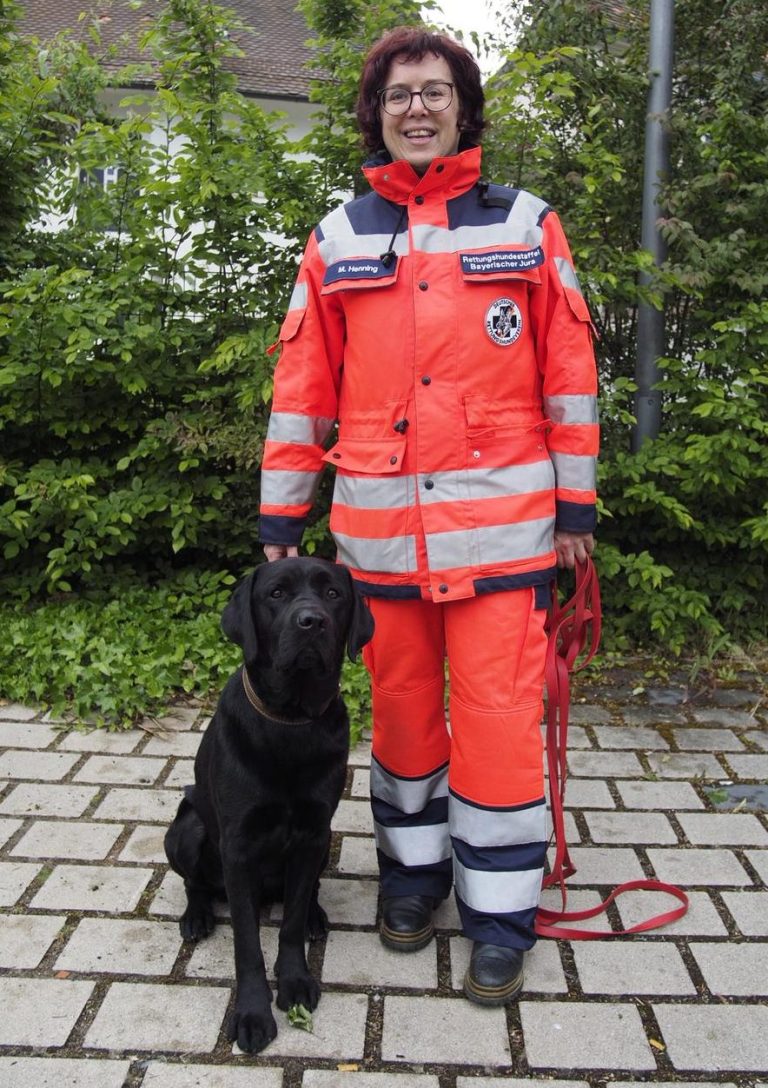 Ehrenamtliches Vereinsmitglied mit roter Weste steht neben einem schwarzen Labrador auf einer Wiese.