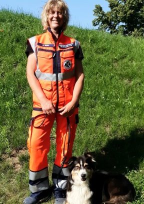 Ein Mitglied im Verein Rettungshundestaffel Bayrischer Jura e. in orangefarbener Einsatzkleidung steht neben einem Mantrailer auf der Wiese.