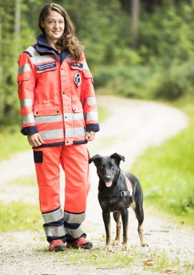 ehrenamtliches Vereinsmitglied (Frau) in orangener Rettungsjacke steht neben einem schwarzem Rettungshund auf einem Waldweg.
