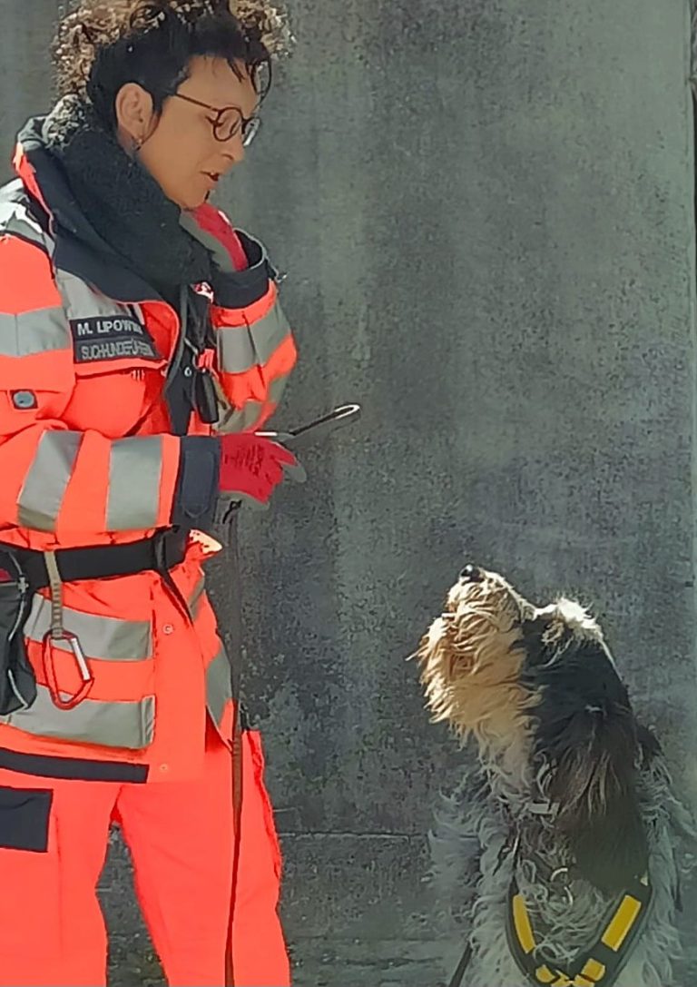 Frau in orangener Rettungsuniform steht mit einem Suchhund an einem Waldweg.