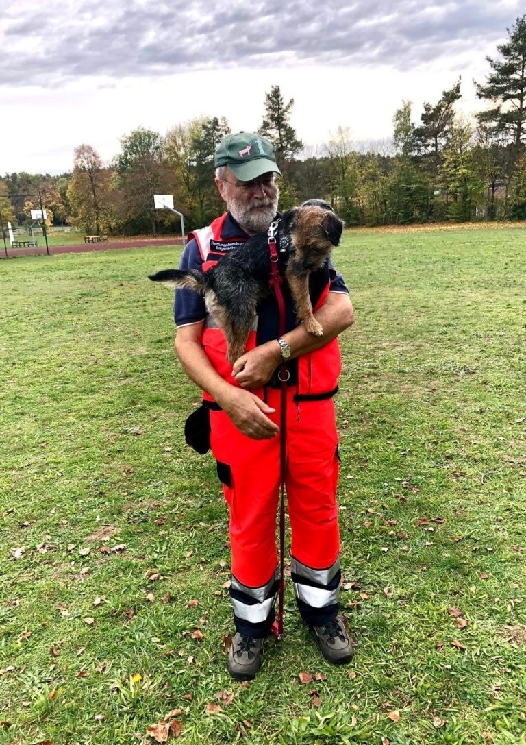 Mann in roter Einsatzkleidung hält einen Mantrailing-Hund auf dem Arm, im Freien auf einer Wiese. 