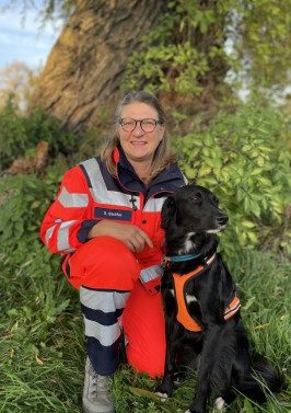 Frau in rot-weißer Warnkleidung sitzt mit einem schwarzen Suchhund in der Natur. 