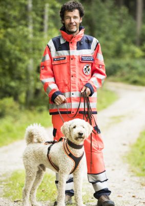 Ein Rettungshundeführer in orangefarbener Kleidung steht mit einem Hund auf einem Waldweg.